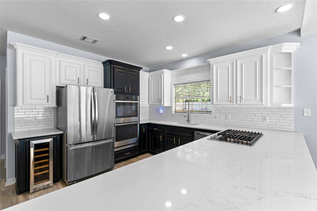 Modern kitchen featuring stainless steel appliances, including a refrigerator, double oven, and wine cooler, with Quartz countertops and a mix of white and black cabinetry. Ample storage with both upper and lower cabinets. The room is well-lit with recessed ceiling lights and natural light.