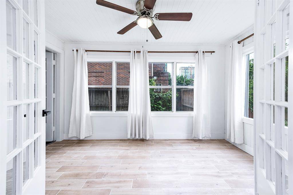 This sunroom features light wood tile flooring, large windows with sheer curtains, and charming French doors opening to the living room. A bright, airy space perfect for relaxing or entertaining.