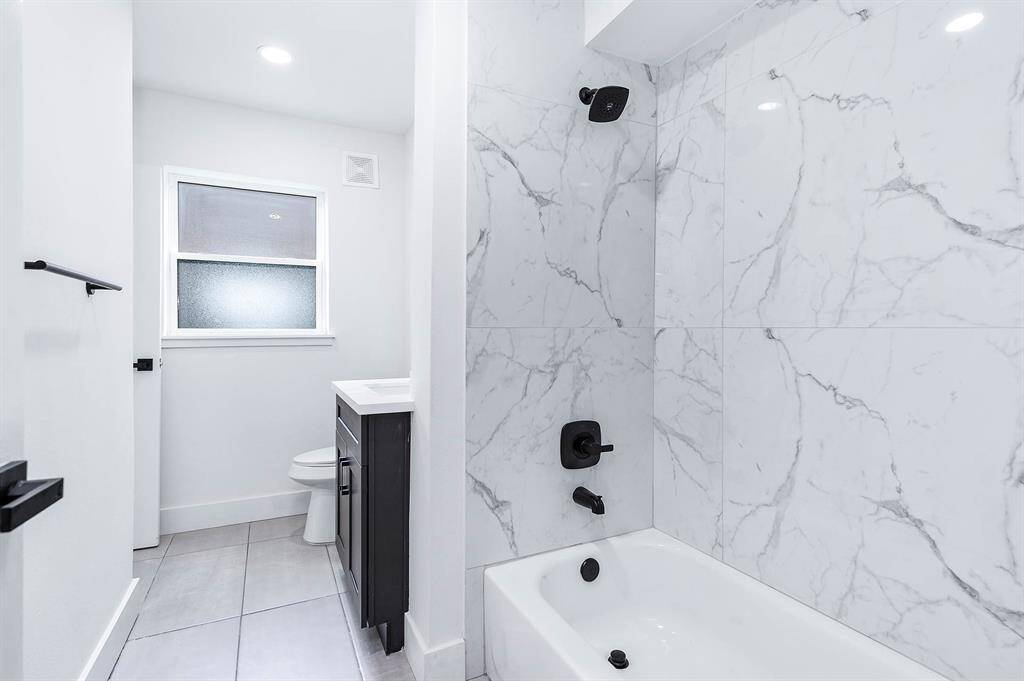 This bathroom showcases a clean, modern design with marble-style tile, a bathtub-shower combo, black fixtures, and a single vanity with dark cabinetry.