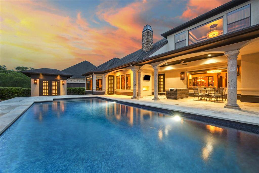 Step into resort-style living with this stunning outdoor retreat, where elegant architectural details meet ultimate comfort. The expansive covered patio features stately columns, ambient lighting, and a cozy fireplace—perfect for evening entertaining. Enjoy al fresco dining beside the sparkling pool, surrounded by lush landscaping and serene privacy. As the sun sets, the glowing sky reflects beautifully off the water, creating a breathtaking ambiance that truly feels like home.