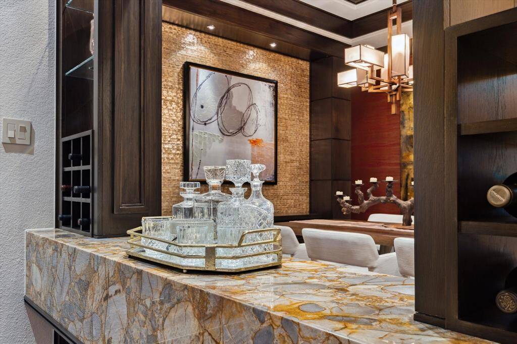 This sophisticated serving bar, seamlessly positioned between the family room and dining area, is a true statement piece. A richly veined imported onyx waterfall countertop anchors the space with luxurious texture and eye-catching depth. Open shelving and built-in wine storage blend form and function, while a backdrop of shimmering tile and bold artwork creates a cohesive visual connection to the adjoining dining room. Designed for effortless entertaining with a hint of glamour.