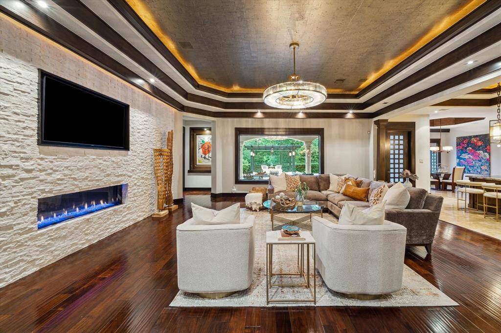 This spacious living room blends comfort and sophistication, featuring a dramatic tiered ceiling with cove lighting and a cork texture ceiling detail. A sleek linear fireplace with crushed marble stacked stone surround adds warmth and ambiance beneath a built-in media display. Rich, hand scraped wood floors flow seamlessly into the open-concept layout, while oversized plate-glass windows frame serene views of the covered outdoor living area and greenspace beyond.
