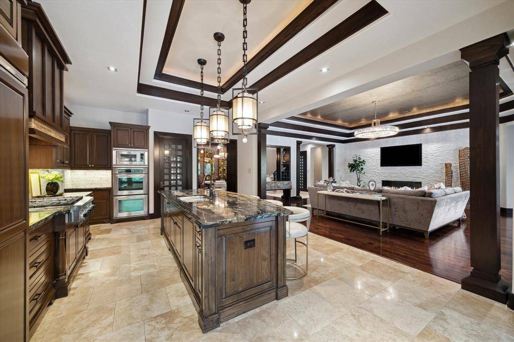 This gourmet kitchen flows effortlessly into the expansive living room, creating a seamless space ideal for everyday comfort. The richly stained cabinetry pairs with exotic granite countertops and a central island that doubles as a roomy gathering spot. Pendant lighting, premium stainless appliances, and tray ceilings with wood accents elevate the overall aesthetic, while the open layout maintains clear sightlines to the fireplace and beyond.