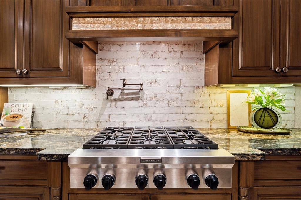A culinary centerpiece, this professional-grade KitchenAid 6–burner gas cooktop is set against a hand-glazed brick backsplash and topped with a custom wood range hood featuring an accent of iridescent mother of pearl tile. The richly veined granite countertops add warmth and sophistication, while under-cabinet lighting and a convenient pot filler elevate both form and function. Every detail is thoughtfully curated for style and performance.