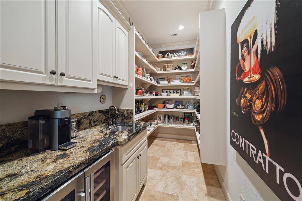 This spacious walk-in pantry goes beyond storage, featuring extensive built-in shelving for organized, easy access to dry goods and serving pieces. A dedicated prep area includes exotic granite countertops, upper soft-close cabinetry, a dual beverage refrigerator, and a built-in sink—perfect for a coffee bar, cocktail setup, or catering support. Tucked just off the kitchen, this space blends practicality with elegance for everyday ease or effortless entertaining.