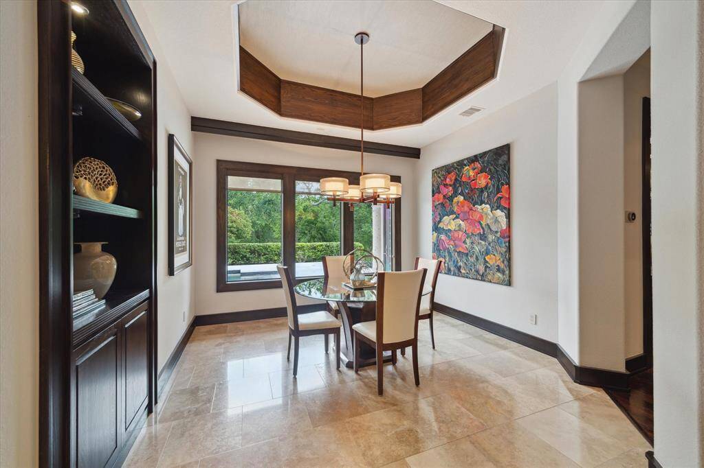 Flooded with natural light, this inviting breakfast room features a deep octagonal tray ceiling trimmed with warm wood accents, designer drum lighting, and polished travertine staggered flooring. A trio of picture windows frames peaceful views of the backyard and pool area, creating the perfect setting for morning coffee or casual meals. A built-in rich wood display cabinet adds elegance and functional storage, while colorful art brings the space to life.