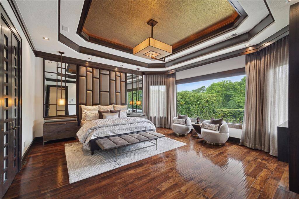 This opulent primary bedroom exudes calm and sophistication, featuring a custom upholstered wall flanked by wood-trimmed mirrors, high-end designer lighting, and a dramatic tray ceiling with inlaid gold leaf accents. A wall of windows covered with lavish draperies frame lush, tree-lined views of the backyard oasis while flooding the space with natural light. The intimate sitting area is ideal for morning coffee or evening unwinding, making this suite the ultimate retreat for rest and relaxation.