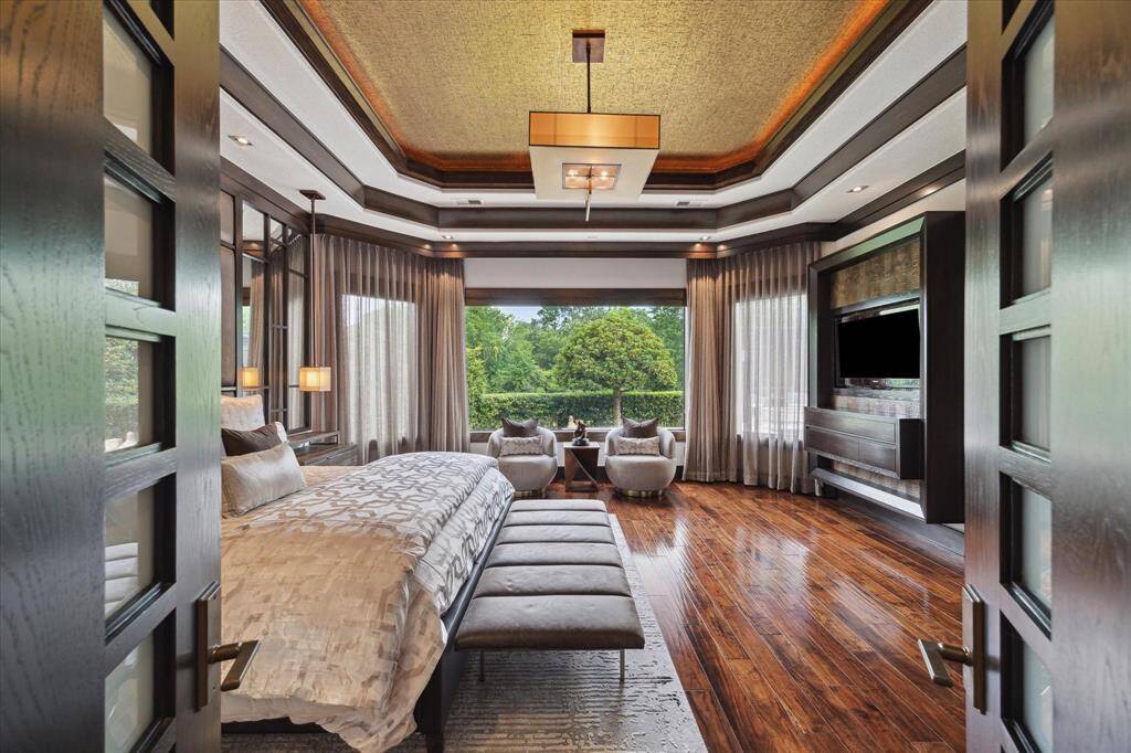 Step through double custom wood and milk glass doors into a refined sanctuary where comfort meets sophistication. Framed by floor-to-ceiling drapery, oversized picture windows bring in tranquil views and soft natural light. The custom tray ceiling with warm metallic inlay and tailored lighting adds architectural interest, while the built-in entertainment center with alligator accents and Musica Net Stream speaker system creates the perfect blend of relaxation and elevated entertainment.
