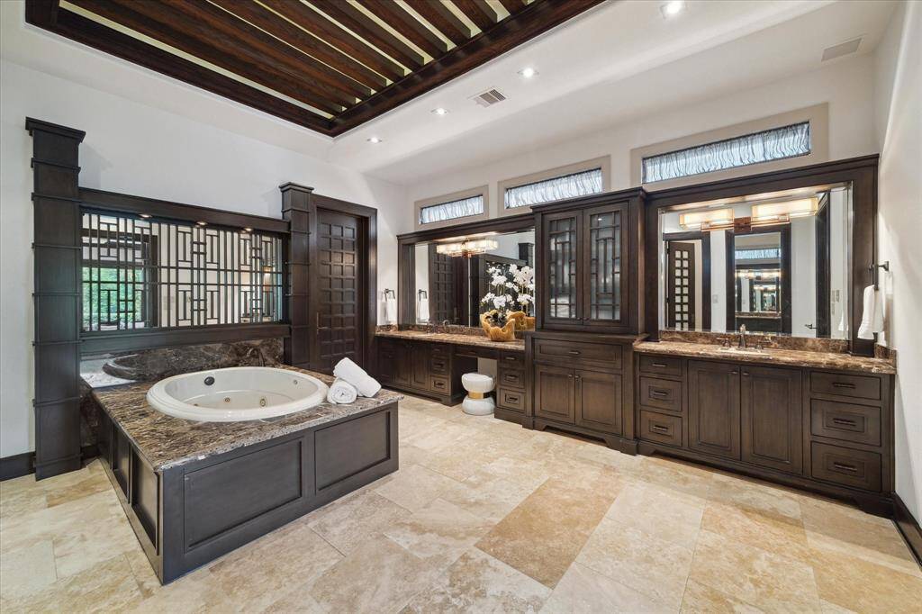 This luxurious primary bathroom offers a true spa-like experience, complete with a deep jetted tub set into a granite-surround platform and flanked by architectural wood columns and a mirrored wall with inlaid wood design.  Dual vanities with built-in linen tower provide ample storage, while upper clerestory windows bathe the room in soft natural light. A dramatic coffered ceiling with lighted exposed beams adds architectural sophistication to this serene space.