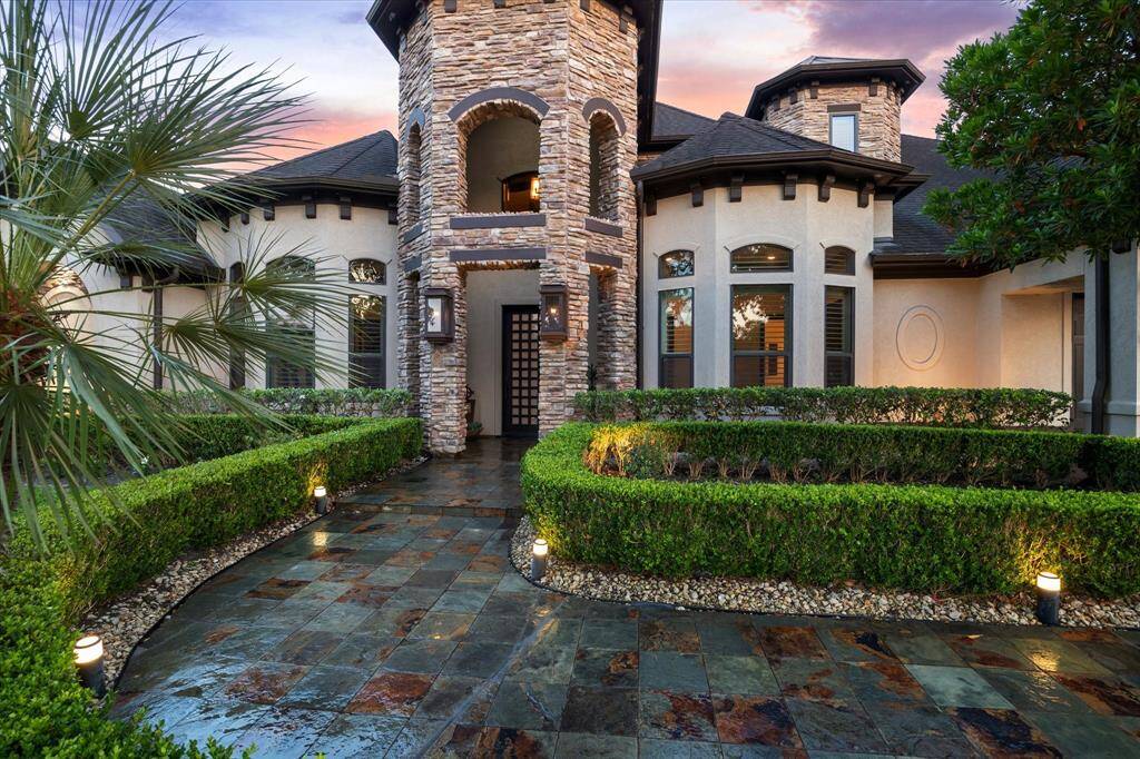 A grand stone turret and arched façade introduce this stunning Mediterranean-inspired estate, where timeless architecture meets refined detail. The slate tile walkway, framed by manicured hedges and soft lighting, leads to a custom wood 8-foot front door with 60 milk glass windowpanes—both elegant and secure. Accented by stacked stone columns and charming gas lantern sconces, this impressive entrance sets the stage for the luxurious living experience inside.