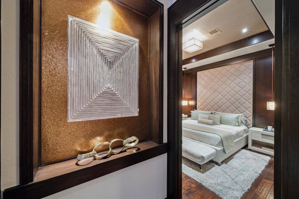 Anchored by an inset art niche featuring a striking illuminated gold leaf backdrop, this stylish transition into the luxurious guest suite sets the tone for refined comfort and relaxation to come. The bedroom beyond features a wood-trimmed coffered ceiling with imported wallpaper inlay, a neutral tufted wall treatment, deep stained wood accent walls, designer pendant lighting, and layered textures that reflect a serene, boutique hotel aesthetic.