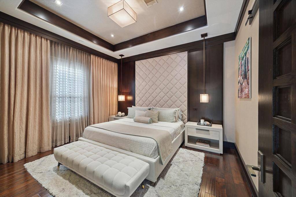 Tucked privately at the rear of the home, this secondary bedroom offers a serene guest retreat complete with a walk-in closet and adjoining bathroom. The tufted wall provides a stylish backdrop for the bed, flanked by deep wood walls and modern pendant lighting. The soft glow filtering through the Plantation shutters and upscale window treatments create a tranquil, retreat-like ambience. Thoughtfully appointed with sophisticated finishes, the suite evokes the elegance of a boutique hotel.
