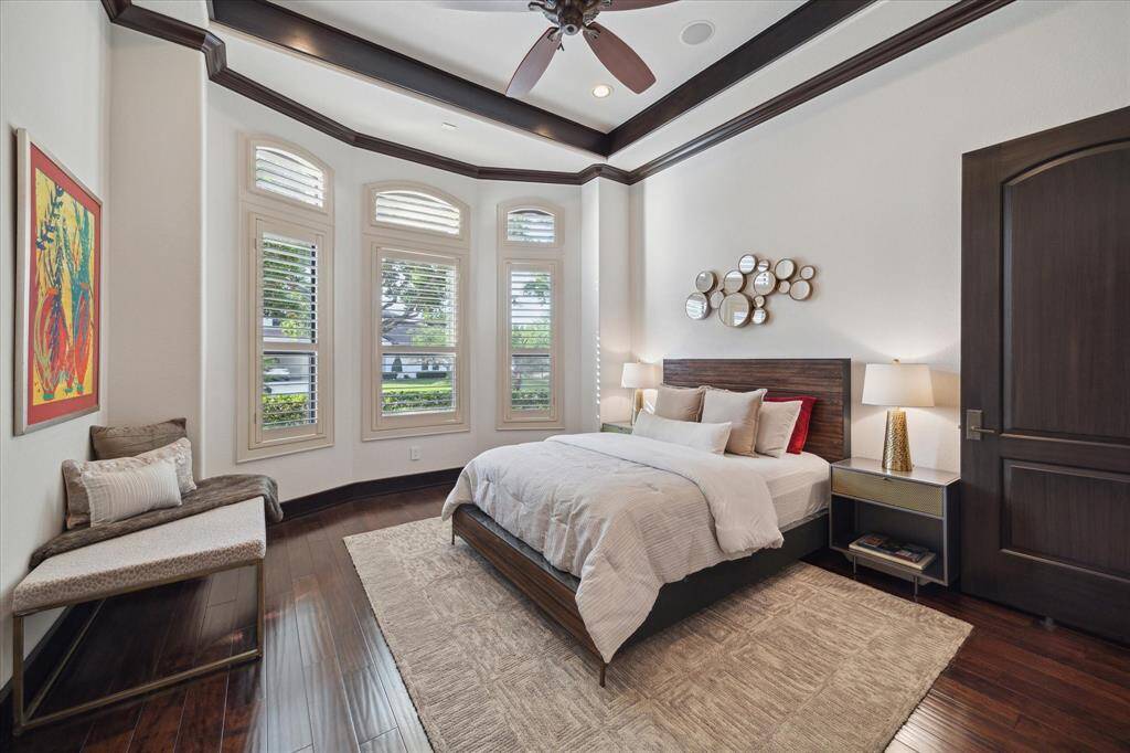 This sunny secondary bedroom enjoys a curved wall of windows with plantation shutters, allowing abundant natural light while maintaining privacy. The clean, modern aesthetic is enhanced by a trayed ceiling with dark wood trim, rich hardwood flooring, and a warm neutral palette. Featuring an ensuite bathroom, walk-in closet with built-ins, and plush textures, this space feels inviting, yet functional—perfect for a guest or family member to unwind in comfort.