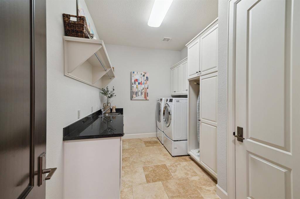 This light-filled laundry room, conveniently located just off the kitchen, is thoughtfully crafted for both efficiency and ease. Abundant cabinetry ensures plenty of storage, while polished granite countertops create a practical surface for folding and organizing. A deep utility sink, integrated hanging rods, and overhead shelving maximize utility, while the neutral tones and travertine flooring lend a crisp, refined finish to this everyday space.