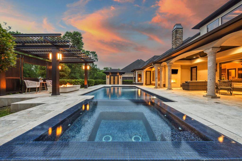 This breathtaking backyard retreat is anchored by a gleaming pool with a deep blue PebbleTec surface and raised infinity spa, accented with deep blue mosaic tile and surrounded by a sprawling travertine patio. At one end, a striking pergola frames a cozy fire pit lounge—perfect for sunset gatherings. Elegant stone columns support the expansive covered patio, complete with an outdoor fireplace and multiple seating areas. Ambient lighting and lush landscaping complete the resort-like ambiance of this private