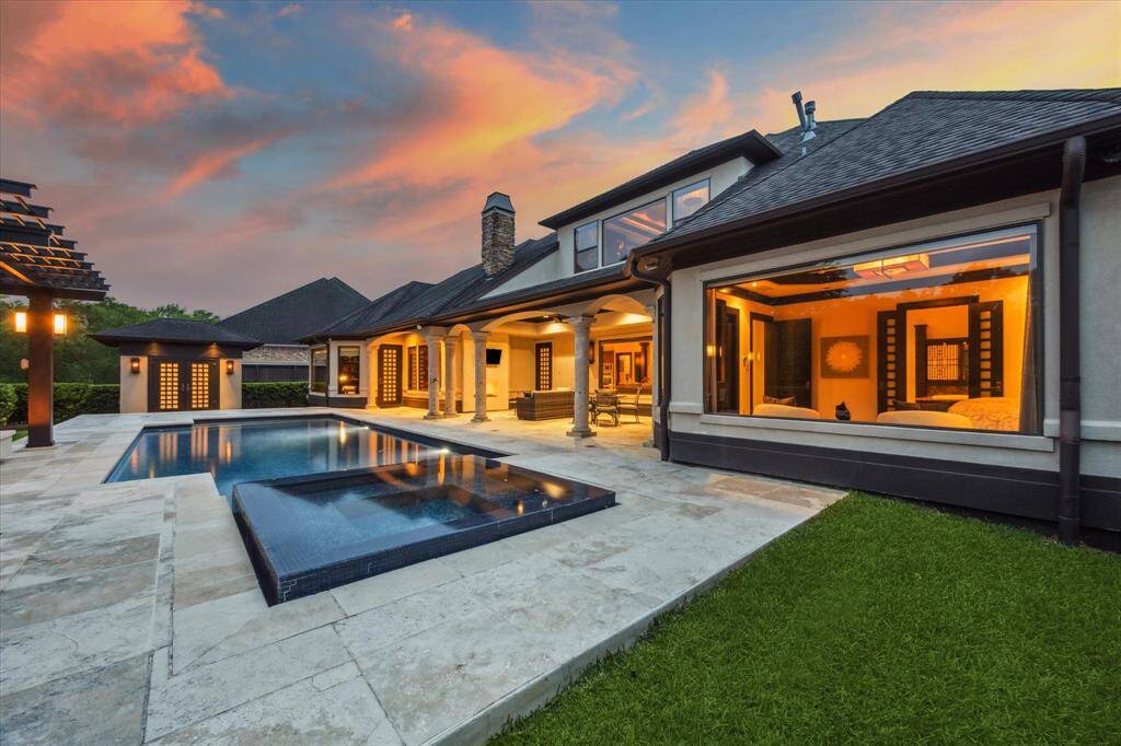 Surrounded by glowing windows and stately stone columns, this twilight view captures the seamless flow between the luxurious indoor living spaces and the serene backyard oasis. The elevated spa spills gently into the sparkling pool, while the surrounding travertine decking enhances the elegant aesthetic. Soft uplighting from the pergola and fire pit lounge creates a magical ambiance, perfect for evening entertaining or quiet relaxation under the stars.