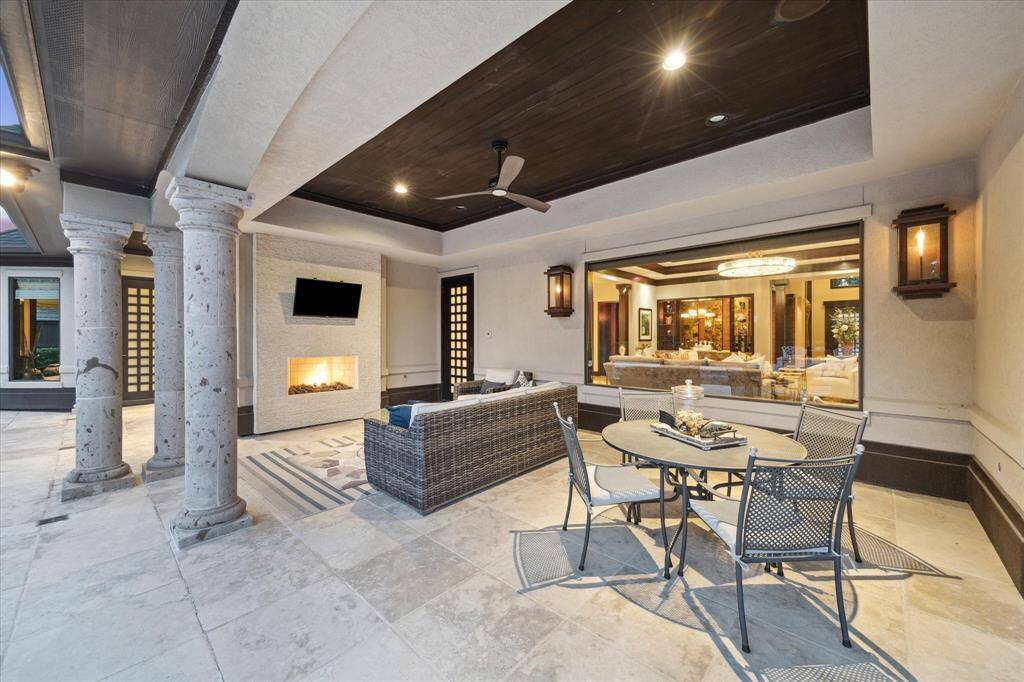 This elegant patio is the ultimate extension of indoor-outdoor living, complete with a gas fireplace with stacked stone surround, gas lanterns and mounted TV for year-round enjoyment. Framed by stately columns and highlighted by a rich wood ceiling with recessed lighting, built-in speakers and a ceiling fan, the space blends sophistication and comfort. Ideal for both lounging and al fresco dining, it’s a seamless connection to the interior entertaining areas just beyond the oversized window.