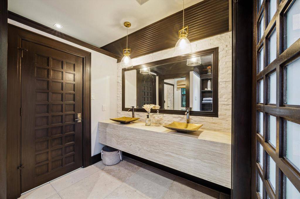 Inside the Zen-inspired air-conditioned pool house, this refined bathroom showcases sleek, spa-like finishes with a floating stone vanity, dual sculptural vessel sinks and striking pendant lighting. A textured stone accent wall adds depth and warmth, while the dark wood trim and custom grid doors maintain a cohesive aesthetic with the home's architectural style. Thoughtfully designed, it offers a luxurious and functional space for post-swim refreshment.