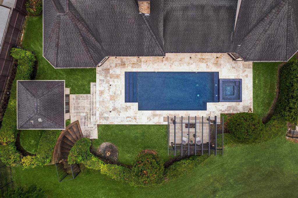 This aerial perspective reveals the meticulously planned backyard oasis, where symmetry and design blend seamlessly. At the heart lies a sleek, rectangular saltwater pool with an integrated spa, bordered by creamy travertine decking that enhances the clean lines of the space. A contemporary pergola with a cozy fire pit anchors one end, while the pool house and gracefully curved staircase to the creek and greenspace beyond create a sense of harmony and balance within the manicured landscape.