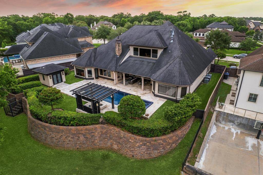 This elevated view captures the full scope of the home's resort-style backyard, artfully enclosed by a curved retaining wall that enhances both privacy and visual appeal. The seamless connection between the main residence, Zen-style pool house, and outdoor entertaining spaces reflects thoughtful design. Manicured hedges, a pergola-covered fire pit lounge, and the sleek pool and spa create a luxurious outdoor living experience in perfect harmony with the natural surroundings.