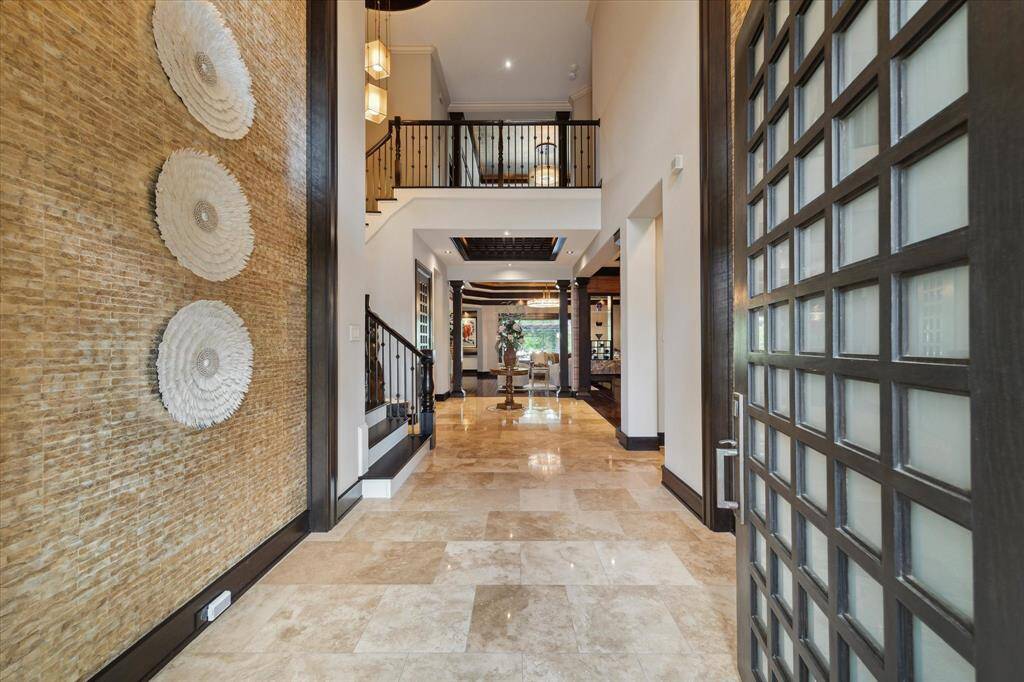Welcome to a showstopping entrance where a soaring two-story foyer is wrapped in shimmering mother of pearl tiles, casting a soft iridescence throughout the space. The dramatic accent walls adorned with artistic décor sets the tone for refined luxury living. Gleaming travertine floors extend through the entry, while the soaring ceiling showcases a sampling of the $250,000 lighting upgrades found throughout the home, which blends grandeur with warmth in this stunning entryway.