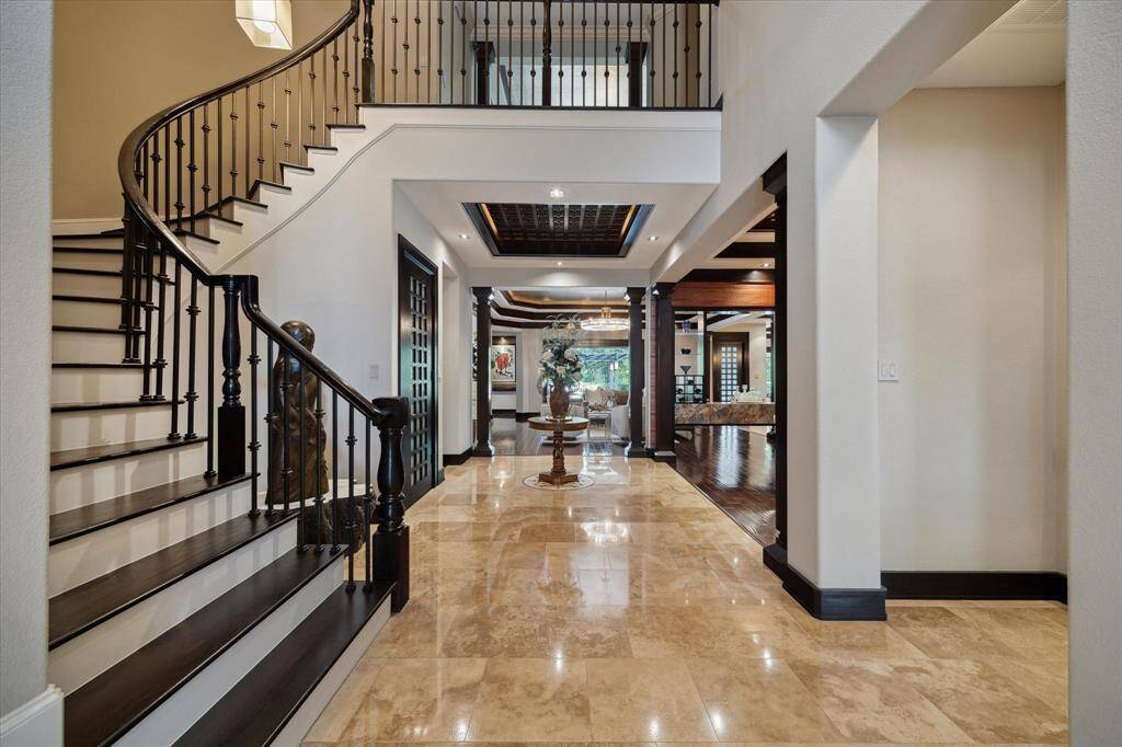 The grand entry flows effortlessly into the heart of the home, where a curving staircase with wrought iron spindles and dark wood accents provides a dramatic architectural centerpiece. Travertine flooring transitions smoothly into wide-plank hardwood, guiding guests over the inlaid mosaic tile floor and through stately columns that define the open layout. The handcrafted wood coffered ceiling lends timeless elegance and architectural interest to the space, beautifully highlighted by ambient lighting.