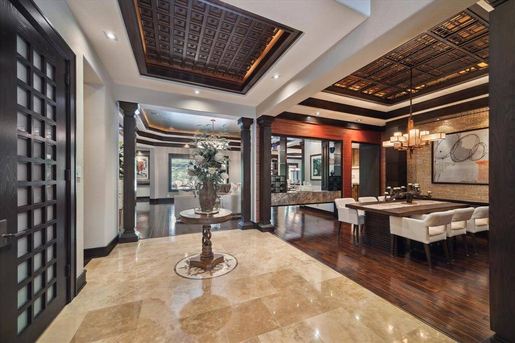 Step through the elegant entryway into this captivating transition space where layered coffered ceilings with intricate custom millwork and ambient lighting create architectural flair overhead, which truly emulates the finest of five-star resorts. Flanked by stately columns and 8-foot custom wood doors with milk glass panes, the open layout effortlessly connects the formal dining room and living areas, all unified by a blend of polished travertine and wide-plank hardwood flooring.