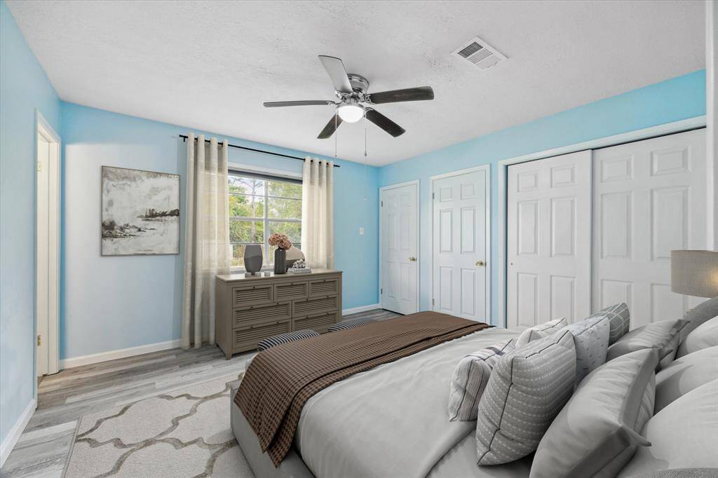 This is a spacious, well-lit room with modern gray wood-look flooring, fresh light blue-gray walls, and a textured ceiling with a ceiling fan. The room features a unique front door with decorative glass, two additional doors, and a window that offers a view of the outside.