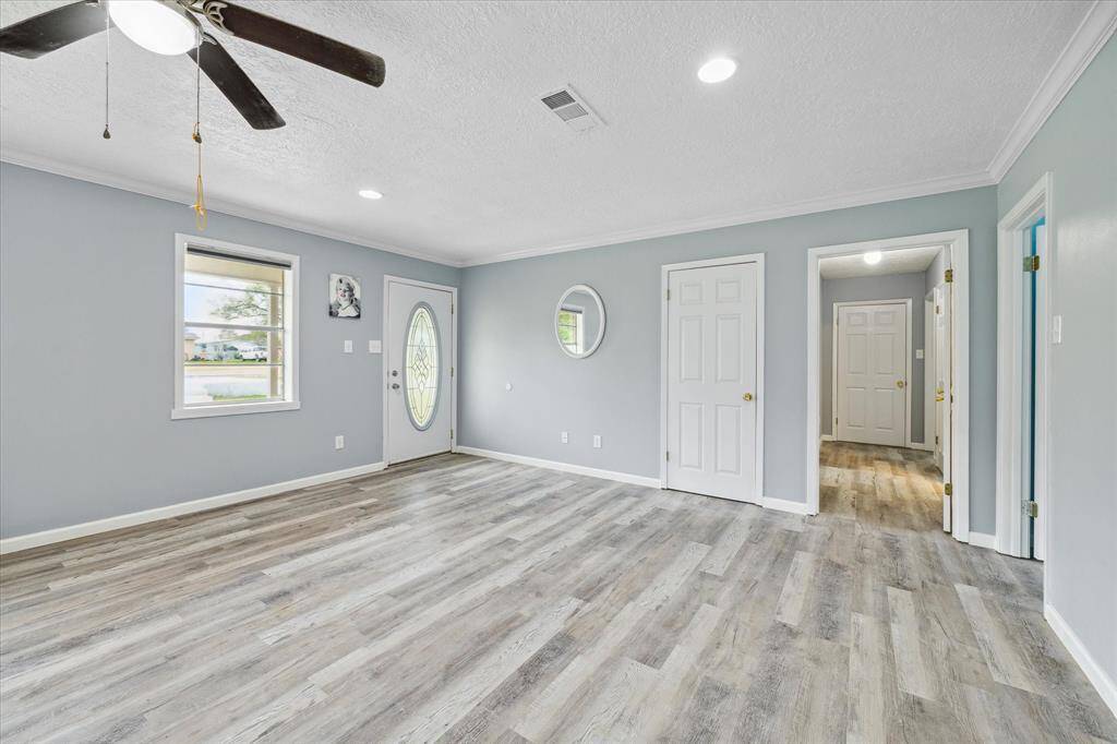 This is a spacious, recently updated room featuring modern, wood-look flooring and a fresh coat of light blue paint on the walls. The room includes a large kitchen island with blue cabinetry and a contrasting gray countertop. Natural light floods in through multiple windows, and there's a ceiling fan for air circulation. The space appears versatile, suitable for a combined kitchen and living area, with potential for ample seating and dining arrangements.