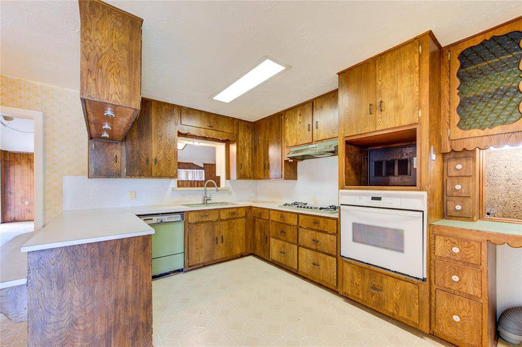 This photo showcases a vintage kitchen featuring wood cabinetry, a built-in oven, and a gas cooktop. The space includes a unique green dishwasher and a pass-through window. There's ample storage and a bright overhead light, adding a retro charm to the room.