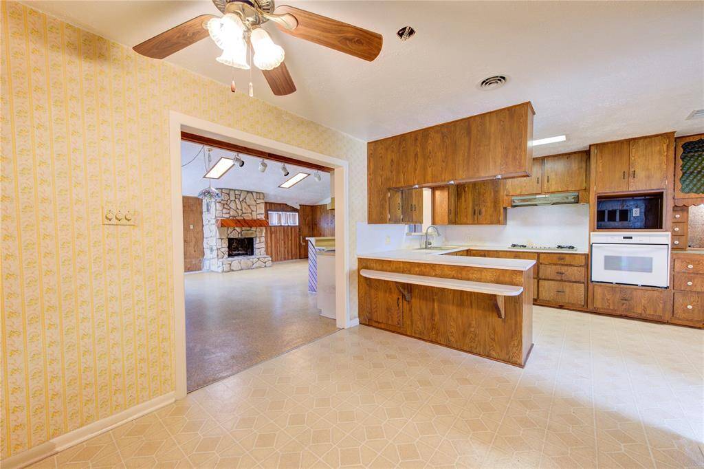 A view of the living room which features a stone fireplace and wood-paneled walls, creating a rustic charm. A ceiling fan and patterned wallpaper add character to the space.