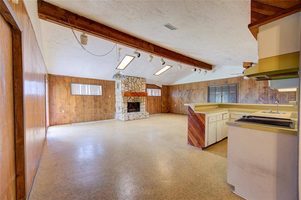 Spacious open-concept living area featuring a stone fireplace, wood-paneled walls, and a vaulted ceiling with exposed beams. The room includes a kitchen with a peninsula for seating and ample counter space, creating a cozy yet functional space for gatherings.