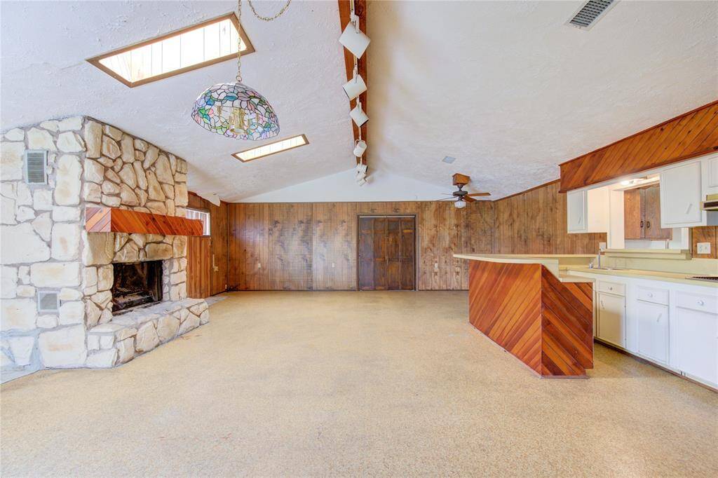 This spacious room features a rustic stone fireplace, wood-paneled walls, and a vaulted ceiling with skylights. It includes an open kitchen with a unique, angled wood design and ample counter space, perfect for entertaining.