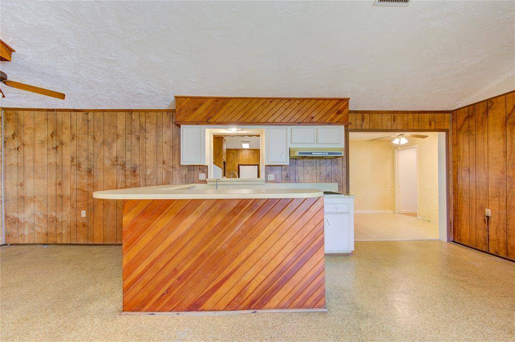 This photo showcases a vintage-style kitchen with wood paneling on the walls and a peninsula counter. The space includes white cabinetry and a ceiling fan, leading into an adjacent room with similar decor.