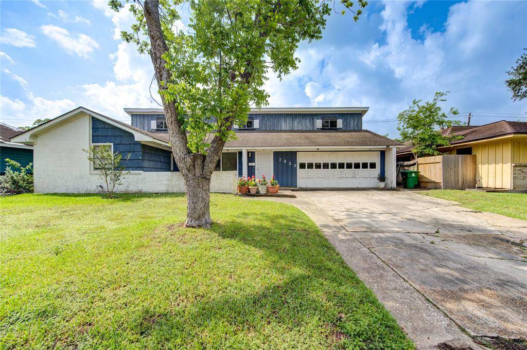 This photo shows a house with a combination of brick and wood siding. It features a garage, a concrete driveway, and a front yard with a large tree. The house has a classic, suburban look with a few potted plants near the entrance.