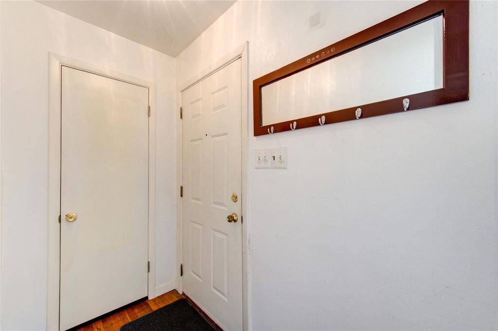 This entryway features a simple, clean design with white walls and a wooden floor. It includes a door with a gold handle, a closet door, and a wall-mounted mirror with hooks for convenience.