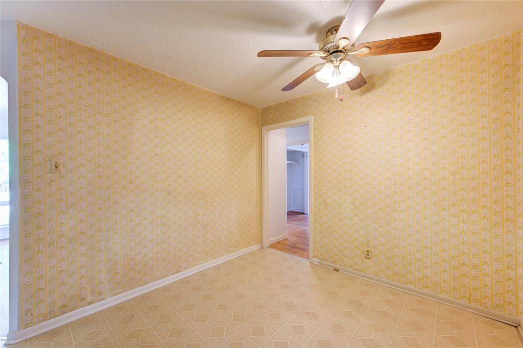 This room features vintage wallpaper and a ceiling fan with wooden blades, with a neutral tile floor leading to another room with hardwood flooring.
