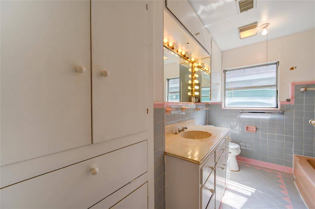 This bathroom features vintage charm with pastel blue and pink tiles, a vanity with ample storage, and classic Hollywood-style mirror lighting. Natural light streams in through a window, enhancing the retro aesthetic.