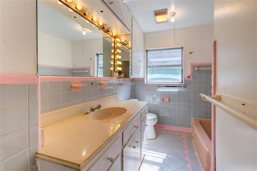 This bathroom features a retro design with pink and gray tile accents, a large mirrored vanity with stylish lights, and a built-in bathtub. It has ample storage and a window for natural light.