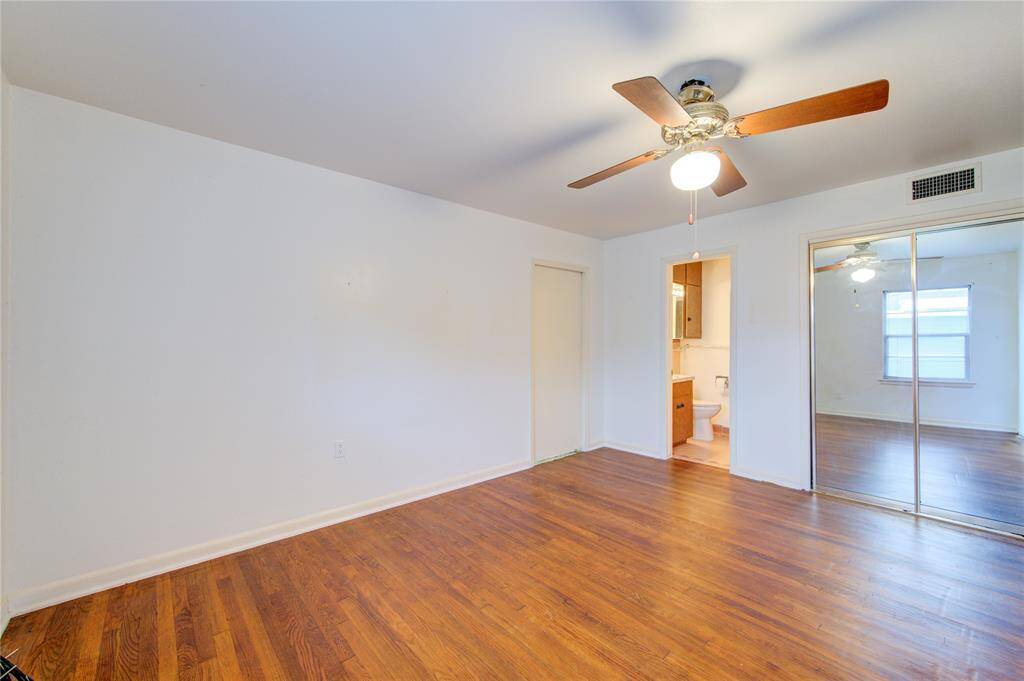 This photo shows a bright bedroom with hardwood floors, a ceiling fan, and a mirrored closet. It features an en-suite bathroom and offers a clean, spacious feel.