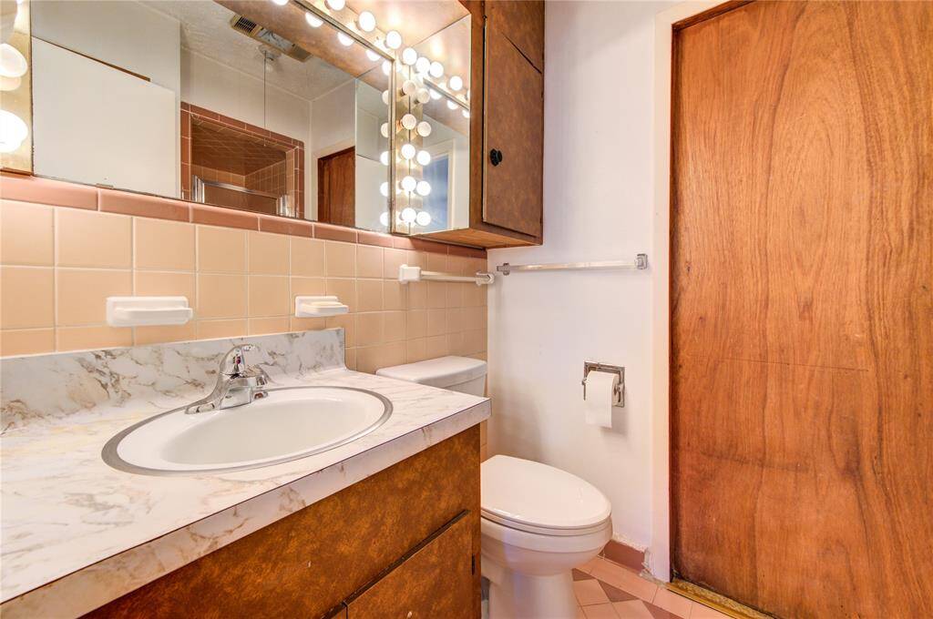 This is a vintage-style bathroom featuring a marble countertop with an oval sink, brown cabinetry, and a large mirror with vanity lights. The walls have beige tiles, and there's a wooden door.