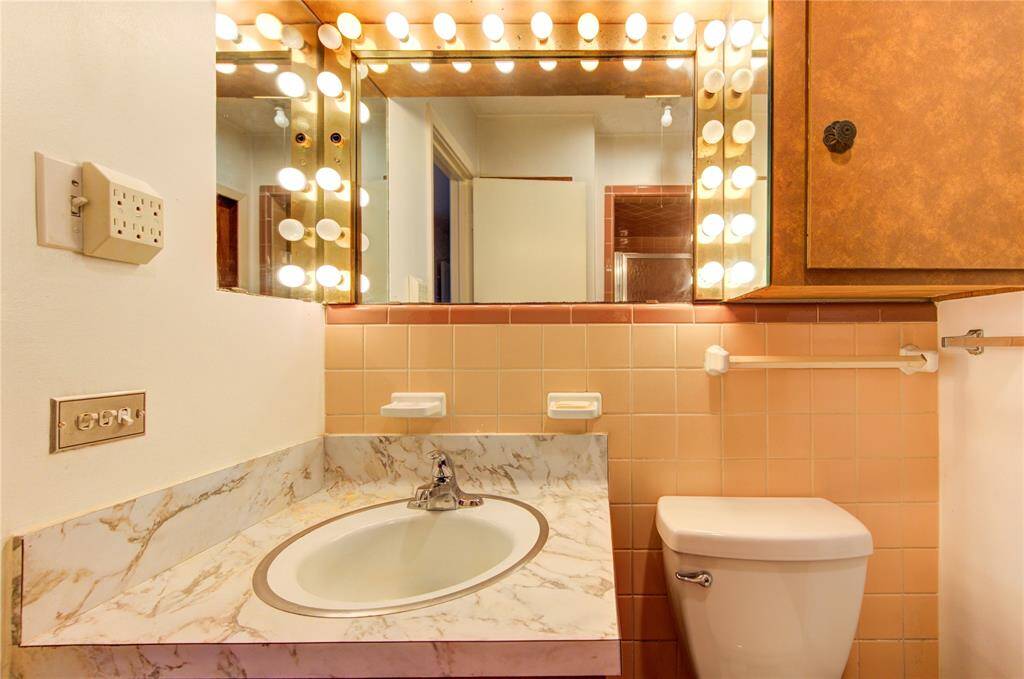 This bathroom features a vintage design with a marble countertop and a round sink. It has a large mirror framed by bright Hollywood-style lights and peach-colored tiles. The space includes a toilet and a wooden cabinet for storage.