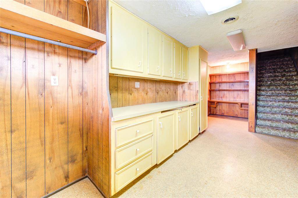This photo shows a utility room with wood paneling, yellow cabinets, and built-in shelves. There's ample storage and counter space, with carpeted stairs leading up. The lighting is bright, enhancing the room's functionality.