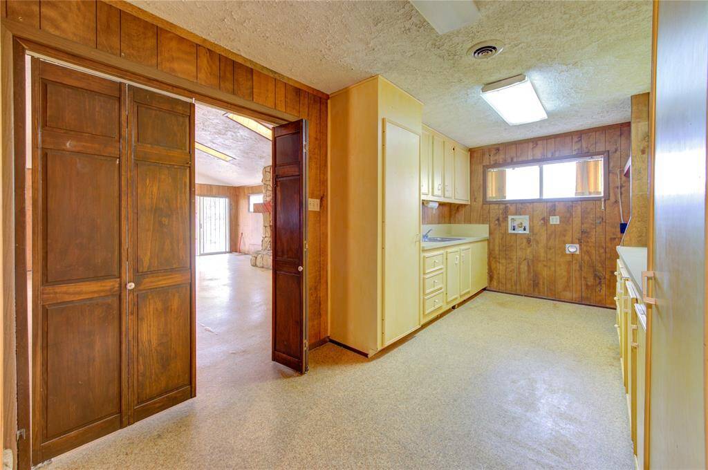 This photo shows a cozy laundry area with wood paneling and ample cabinetry for storage. It connects to a spacious room with natural light coming through large windows, featuring a stone accent wall.