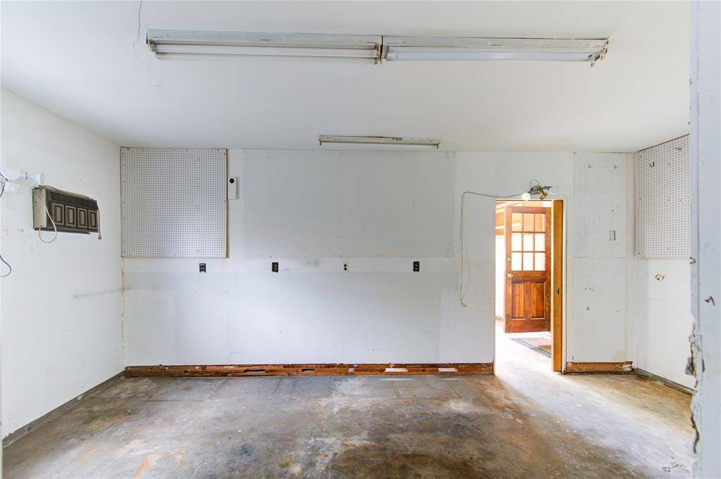 This photo shows a garage space with white walls, pegboards for organization, and overhead fluorescent lighting. The concrete floor and visible door leading outside suggest a functional area for storage or workshop use.
