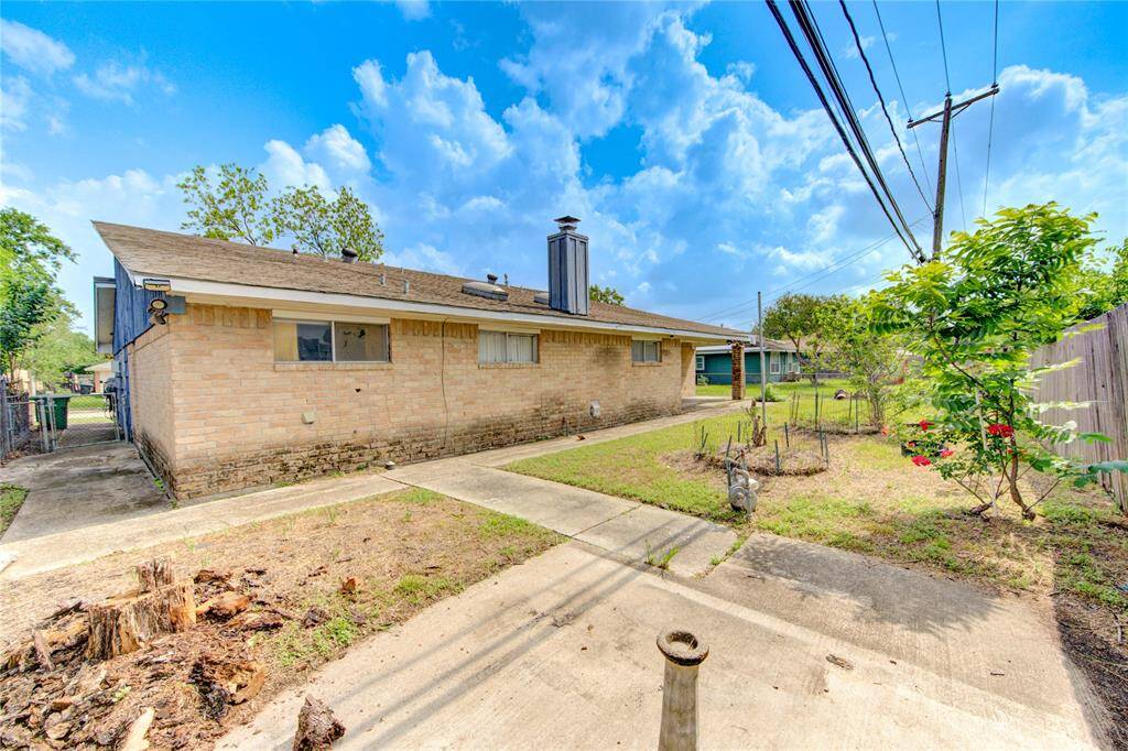 This photo shows the backyard of a single-story brick home, featuring a concrete pathway and small garden areas. The yard is partially fenced and basking in the sunshine of a clear blue sky.