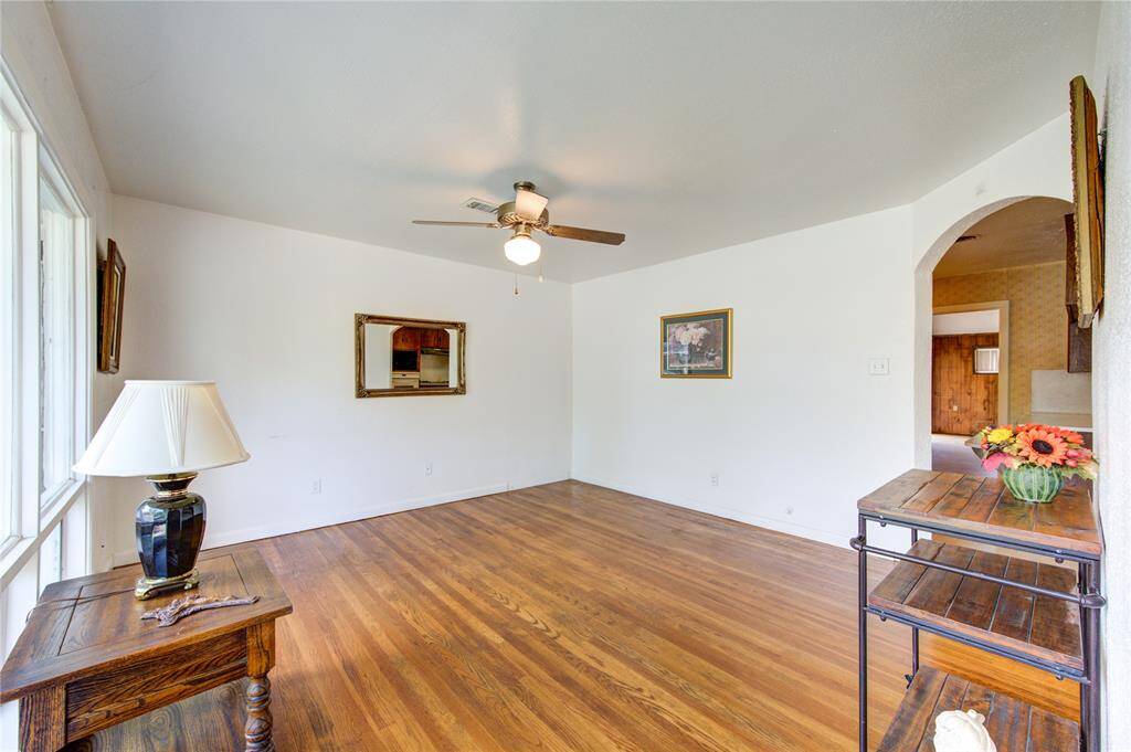 Bright living space with hardwood floors, a ceiling fan, and ample natural light from large windows. The room features neutral walls, decorative wall art, and a cozy archway leading to another area of the home. Ideal for a comfortable and inviting living area.
