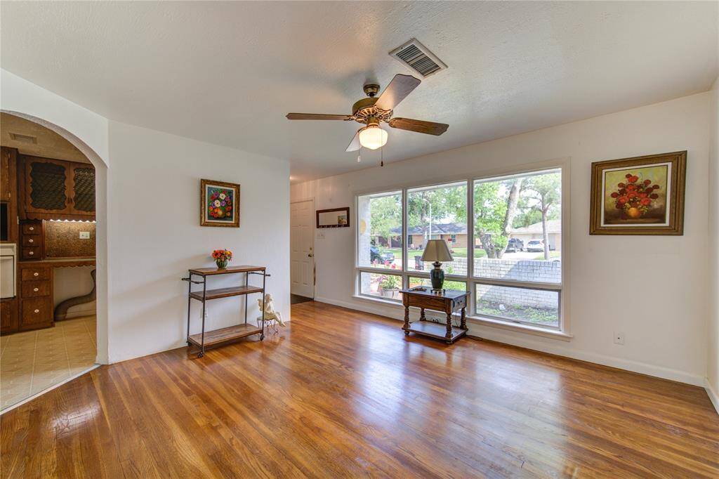 Bright living room with hardwood floors and large windows offering plenty of natural light. Features a ceiling fan and simple decor. An arched doorway leads to the adjacent kitchen area.