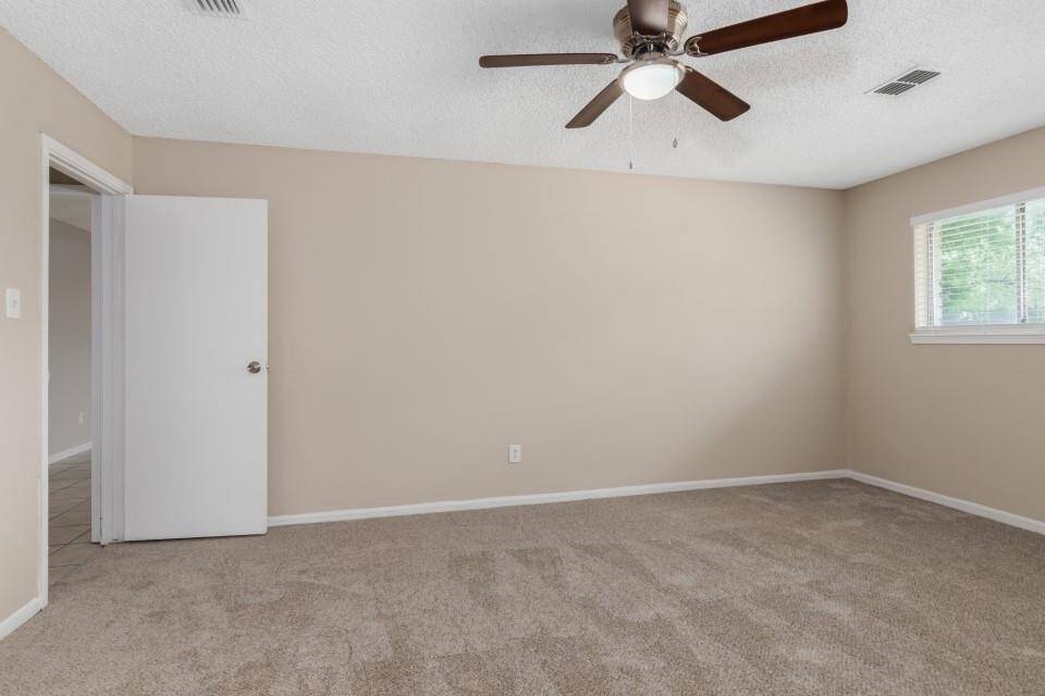 This room features neutral-toned walls and carpet, a ceiling fan with lights, and a window for natural light. It offers a spacious layout ideal for a bedroom or office.