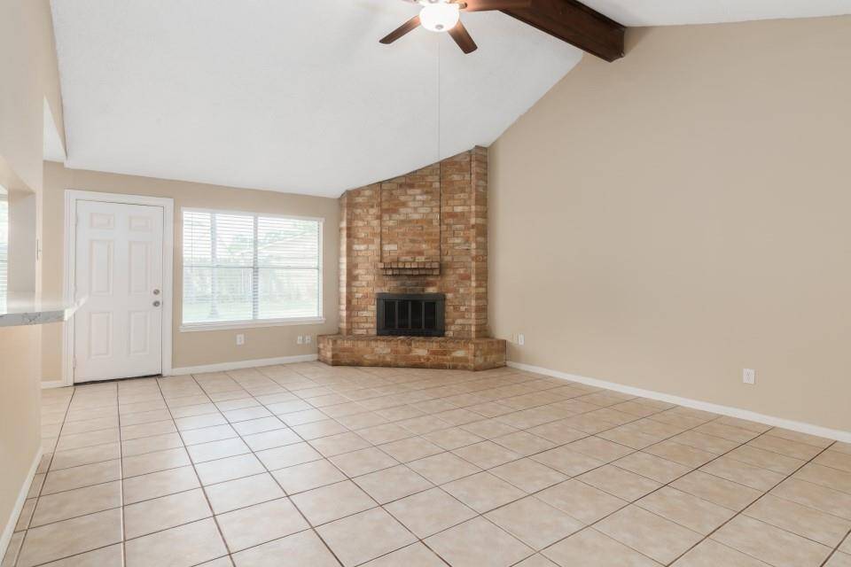 This spacious living room features a vaulted ceiling with a wooden beam, a brick fireplace, and large windows for natural light. It has neutral walls and tiled flooring, creating a bright and airy atmosphere.